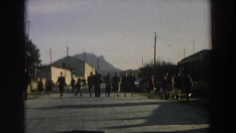 Several people stand or move across a sunlit Italian road while the car approaches, with buildings and a mountain behind them.