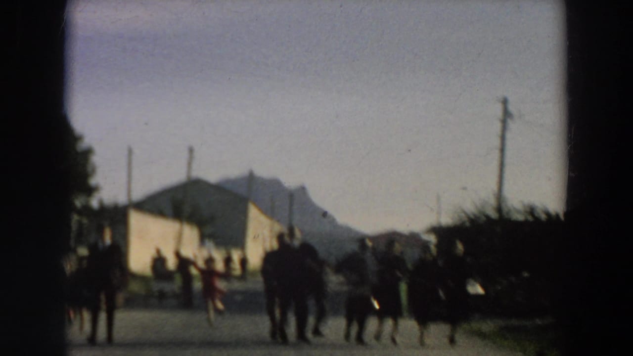 Near the end of the clip, people stand and walk along the road beside a pale building, with a mountain visible in the distance.