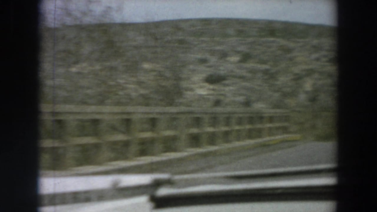 Forward passenger view on a narrow Italian countryside road, with a bridge railing, rocky hillside, and car interior edge visible.