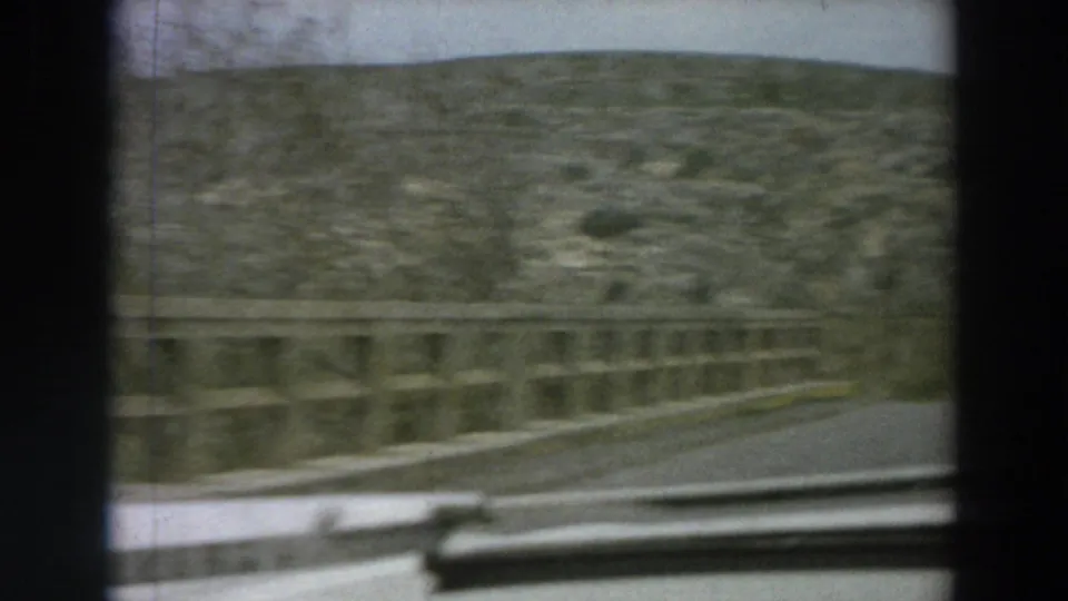 Forward passenger view on a narrow Italian countryside road, with a bridge railing, rocky hillside, and car interior edge visible.