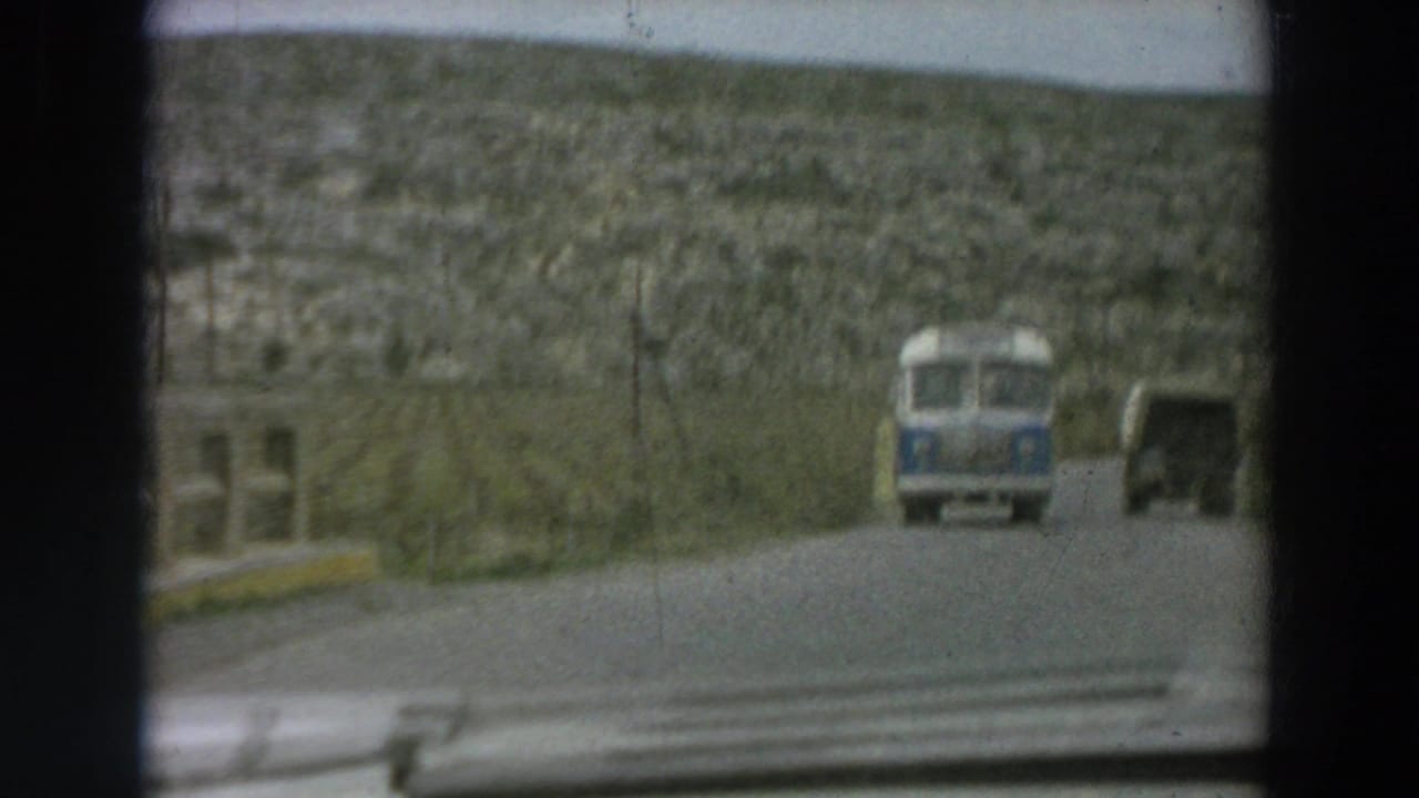 Forward view from the car on a rural Italian road, with a small blue vehicle and trailer ahead against a rocky hillside.