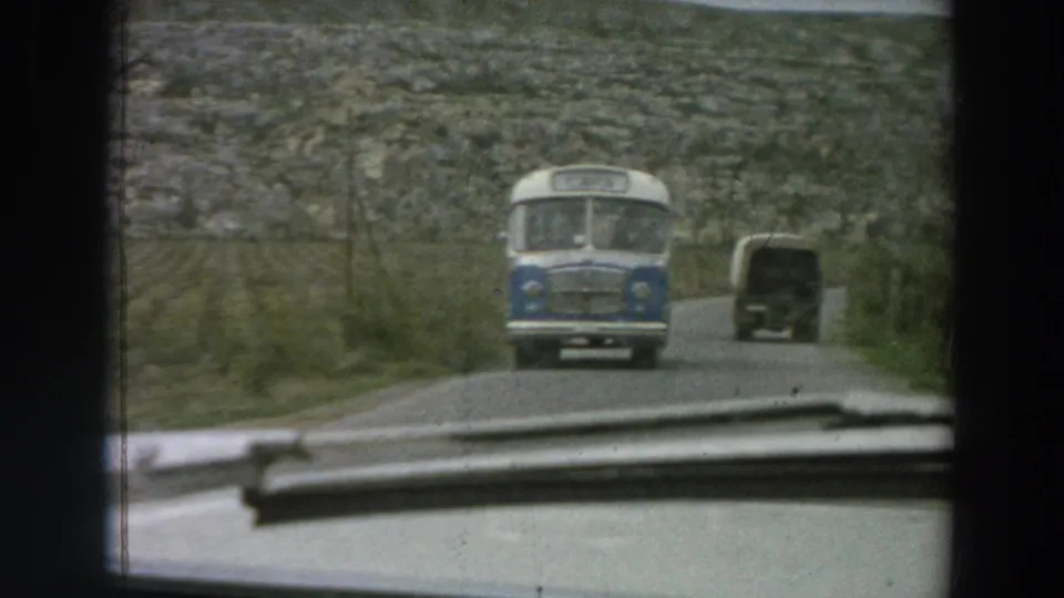 In 1958 Italy, a blue-and-white bus approaches on a narrow rural road, filmed from inside or just behind a moving car windshield.