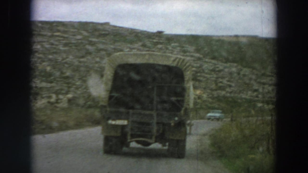 The rear of a dark canvas-covered truck dominates the view as it travels along a narrow road in rural Italy.