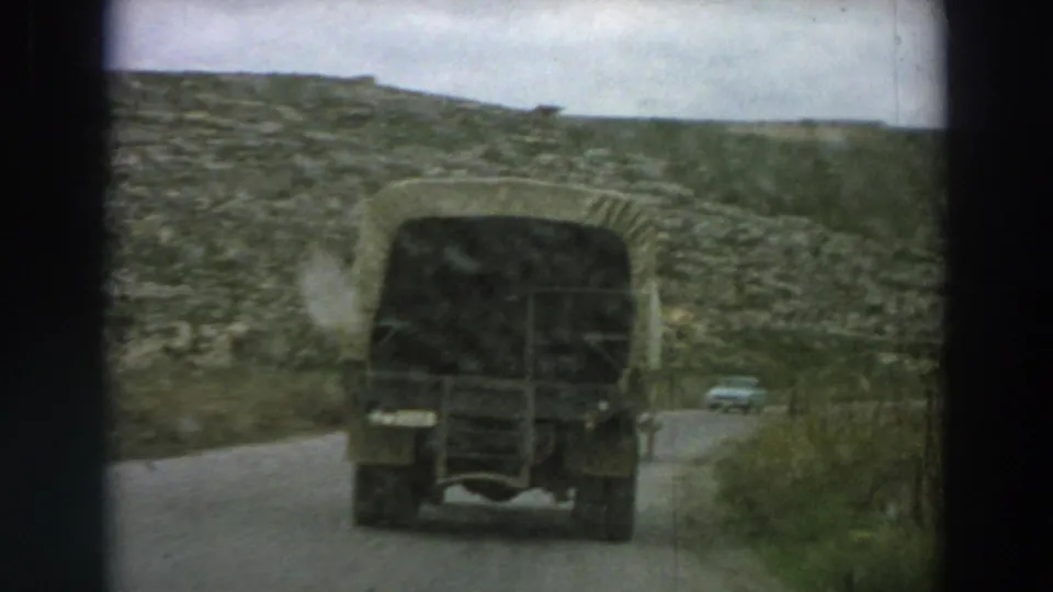 The rear of a dark canvas-covered truck dominates the view as it travels along a narrow road in rural Italy.