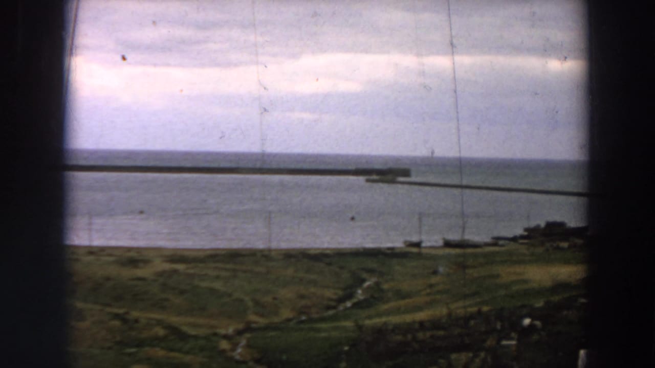 1958 Italy home-movie view of calm sea, grassy shore, and a long pier framed by a dark window or structure.