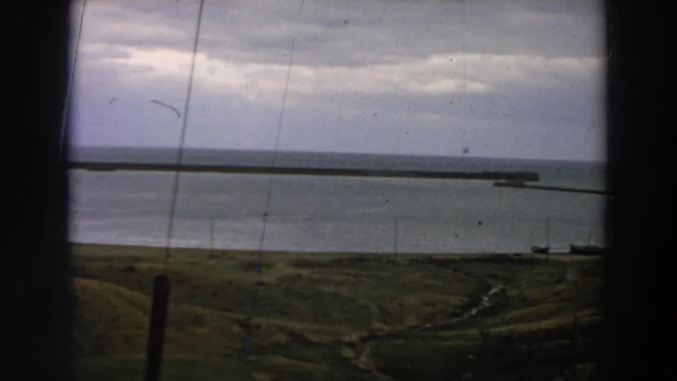 A 1958 Italy coastal harbor view shows calm water, a long pier, shoreline grass, and dark framing in the foreground.
