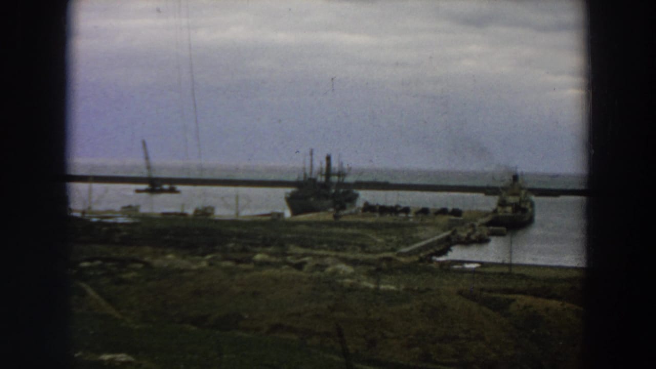 1958 Italy 8mm footage shows two large vessels near a long pier, with grassy harbor land and calm water below.
