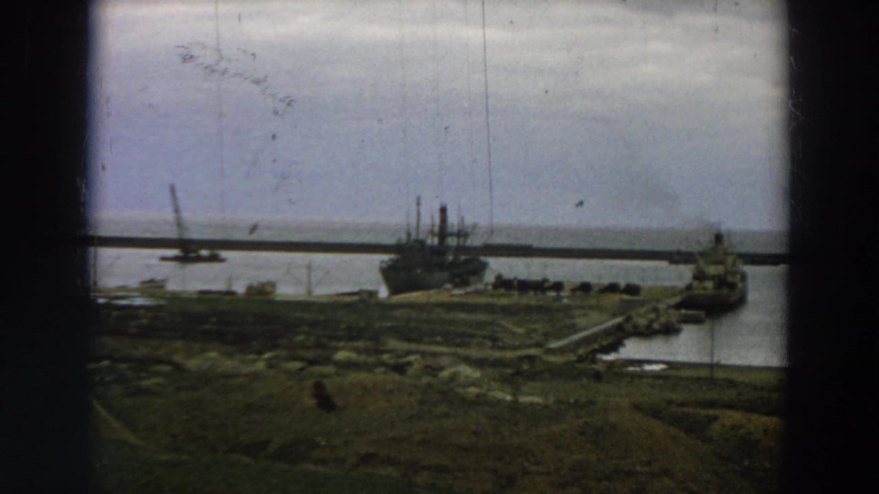 A muted 1958 Italy shipyard view shows anchored vessels beside a long pier, with grassy shoreline and cloudy sky.