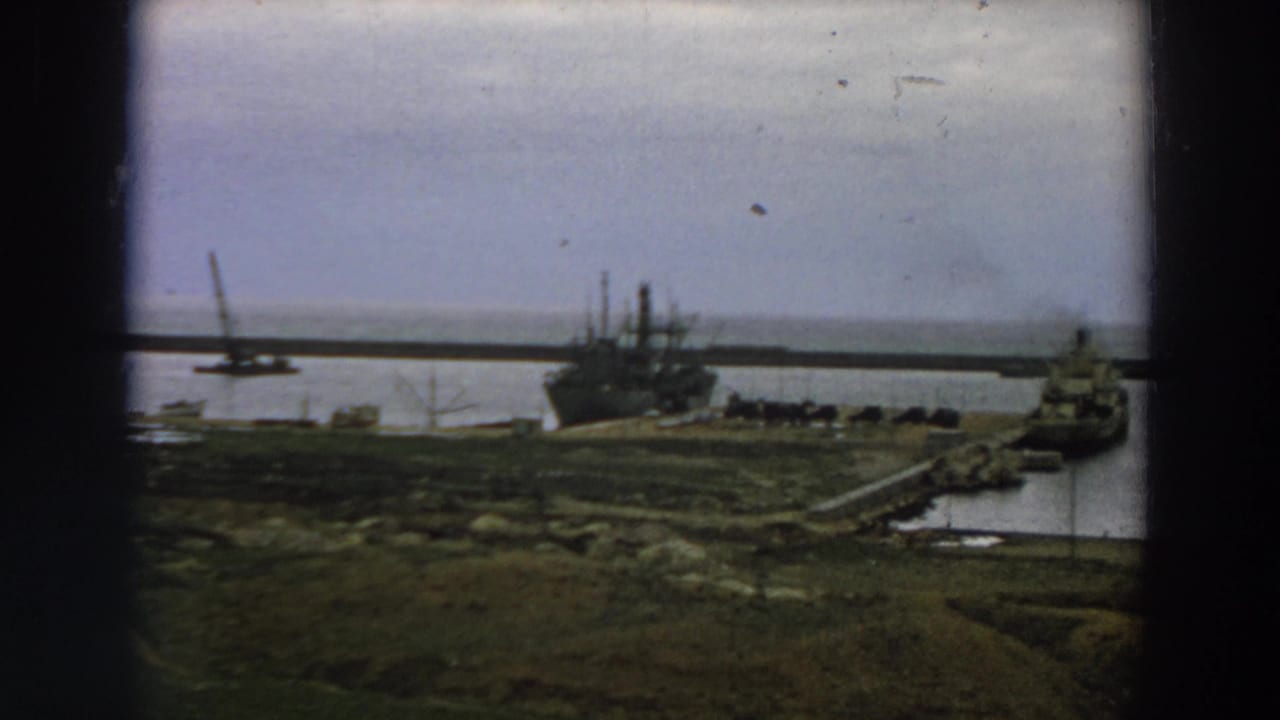 1958 Italy 8mm frame showing a distant moored ship near a pier, low shoreline, water, and a crane in the background.