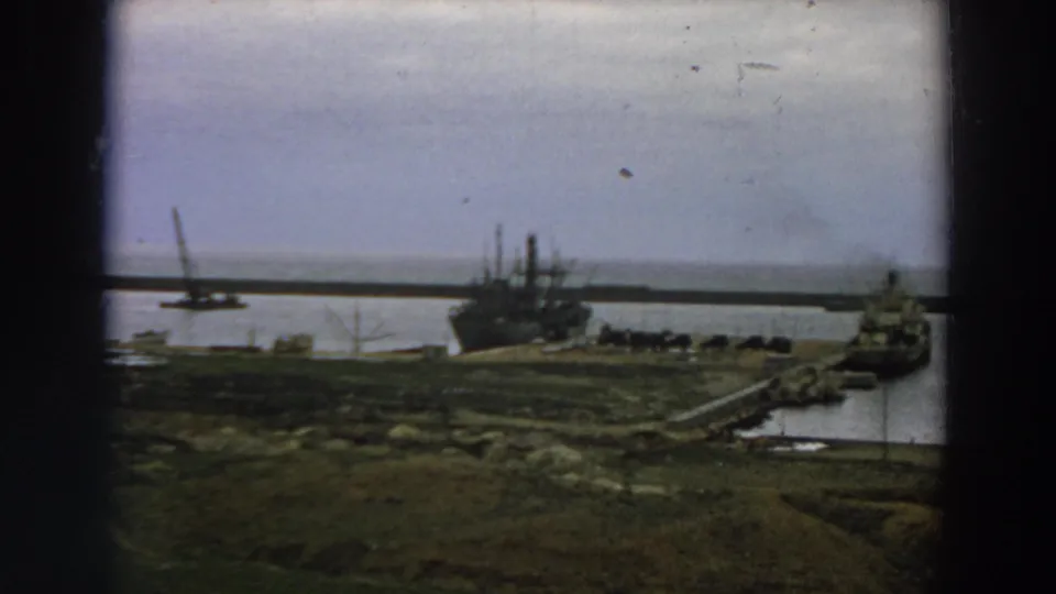 1958 Italy 8mm frame showing a distant moored ship near a pier, low shoreline, water, and a crane in the background.