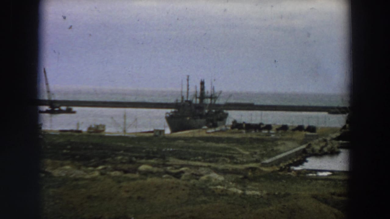 Wide harbor view with a large ship at the pier, water behind it, and rough shoreline in front.