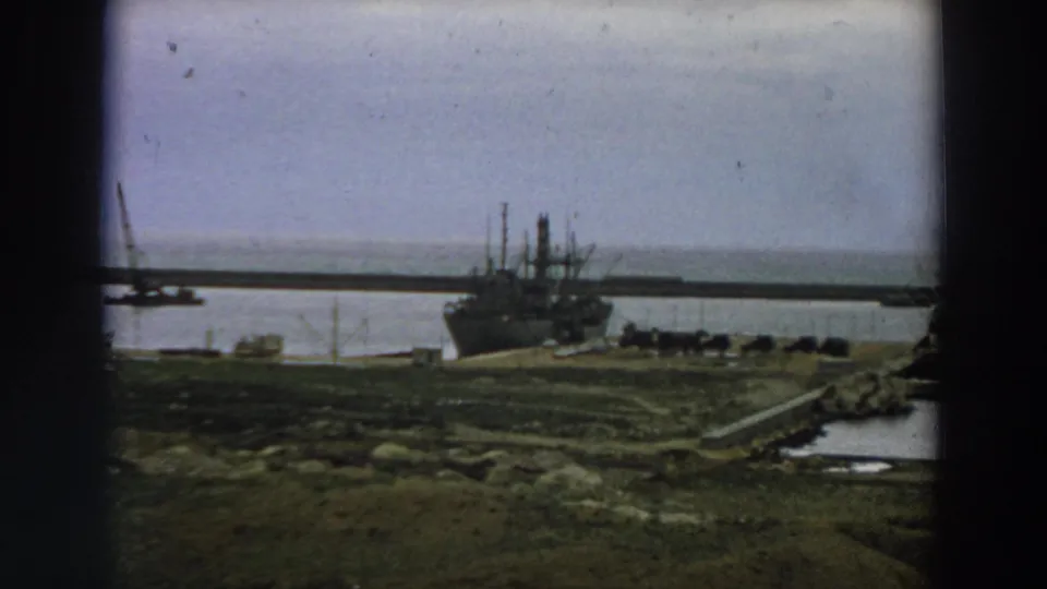 Wide 1958 Italy home-movie view of a large vessel moored at a pier, with water, breakwater, and uneven shoreline visible.