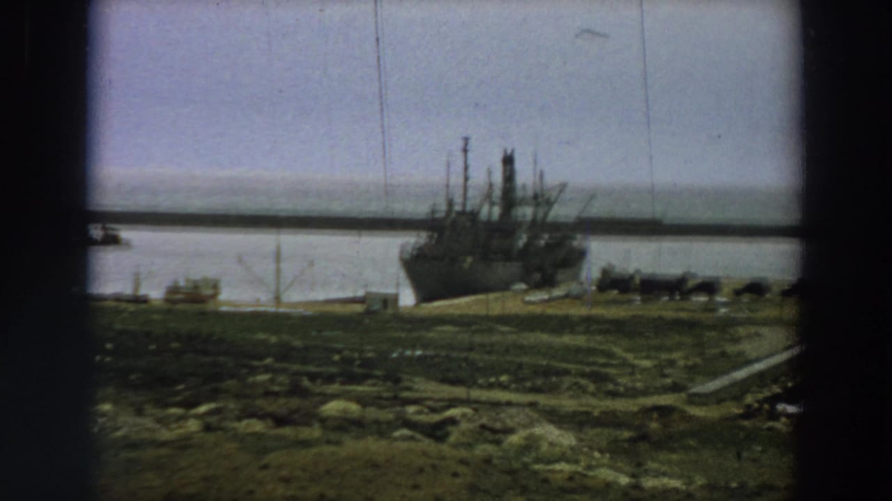 Closer 1958 Italy frame of the moored ship’s bow and pier area, with muted water and rough grass in the foreground.