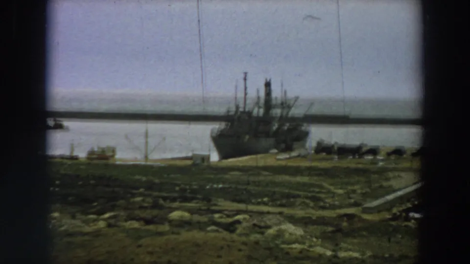 Closer 1958 Italy frame of the moored ship’s bow and pier area, with muted water and rough grass in the foreground.