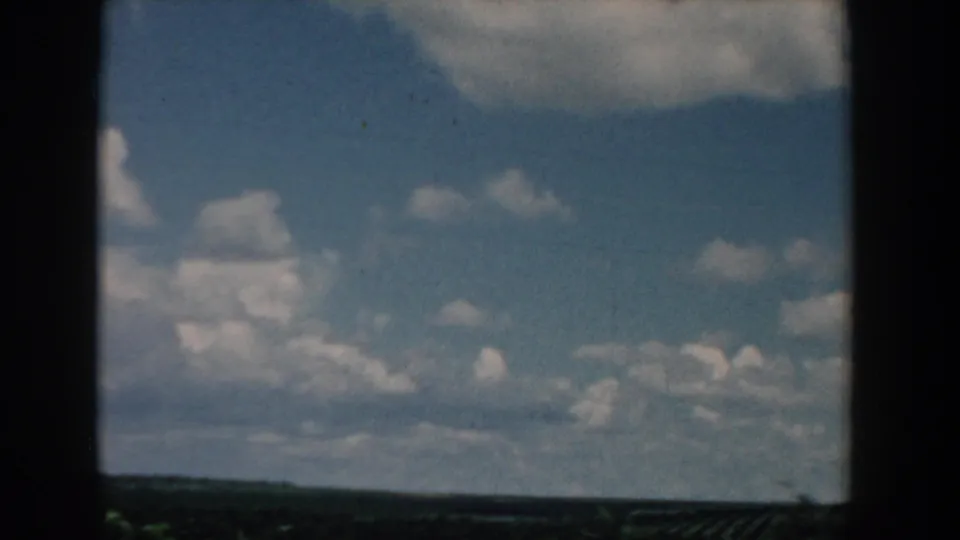 A moving 1958 South Carolina countryside view with large cumulus clouds over a low field horizon and dark foreground edge.