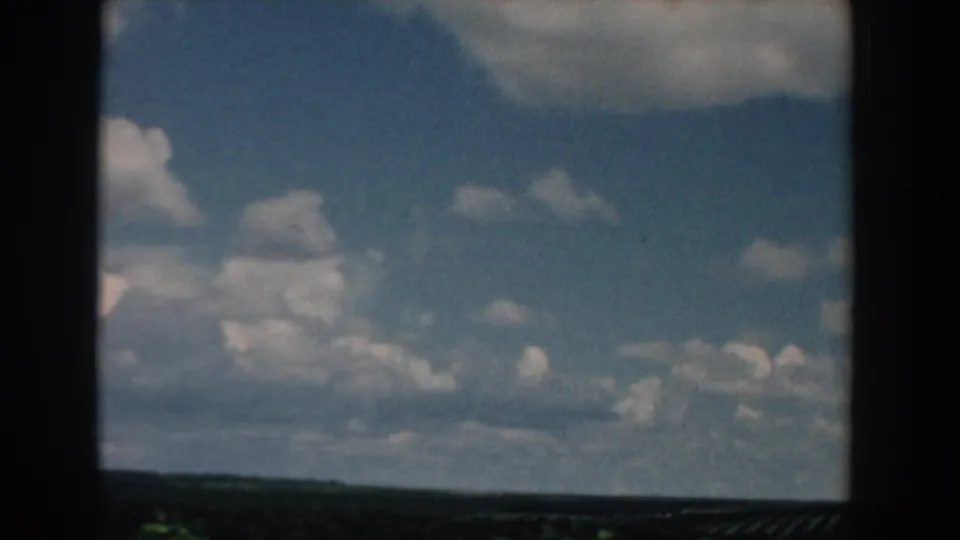 1958 South Carolina home-movie still with cloudy blue sky and distant flat fields, softened by 8mm grain and vehicle motion.