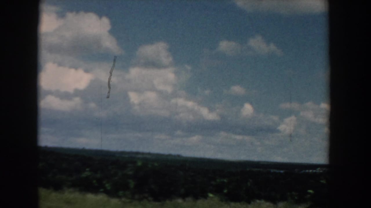 1958 South Carolina home-movie frame showing open green countryside beneath a blue sky with large white clouds.