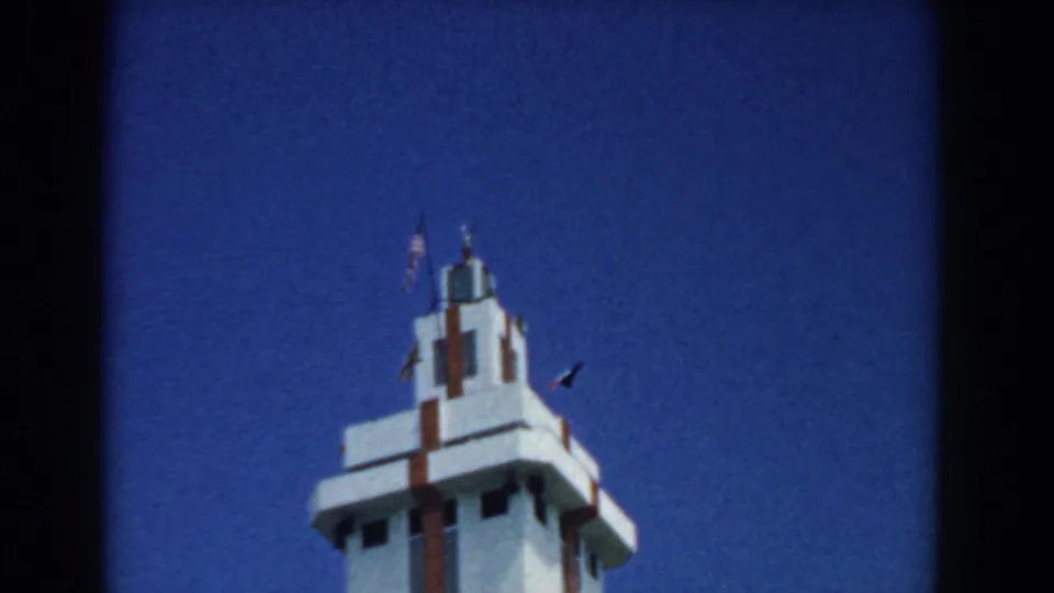 Low-angle home-movie frame of the white South Carolina monument or tower, showing red trim and flags at the top.
