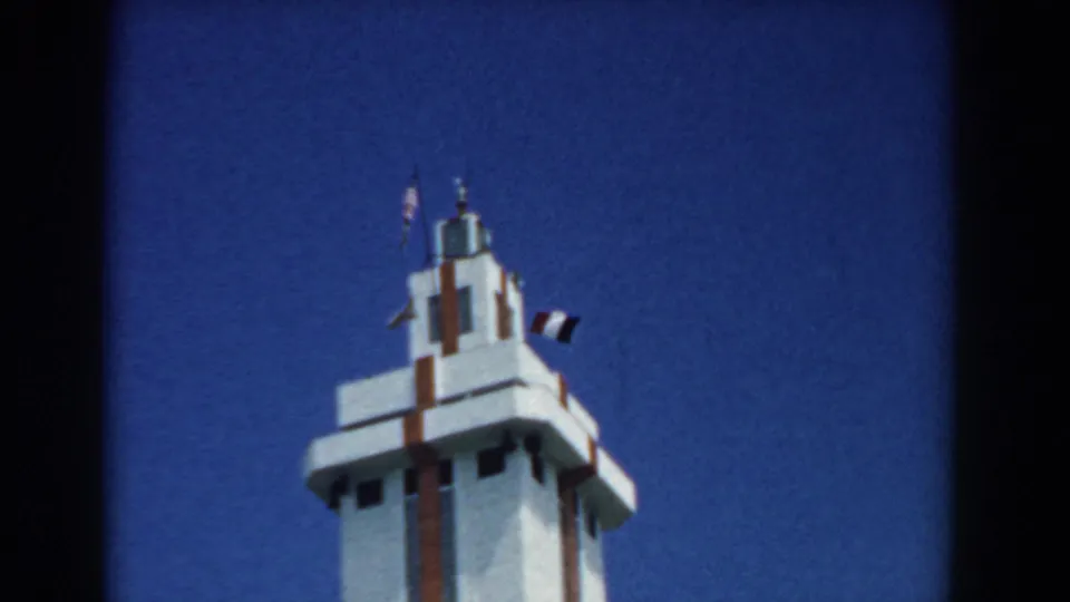 1958 South Carolina 8mm frame showing the upper section of a white tower with red trim and flags flying in a blue sky.
