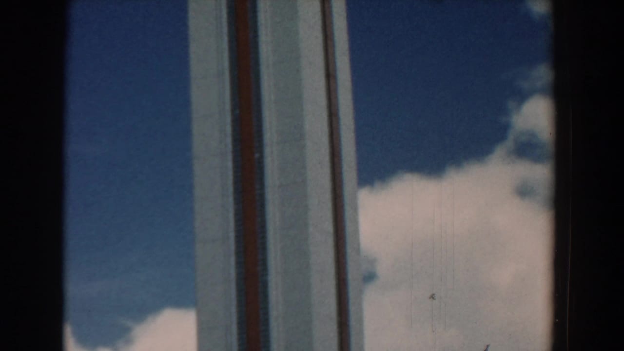 Tall building facade seen through dark window framing with blue sky and clouds behind it.