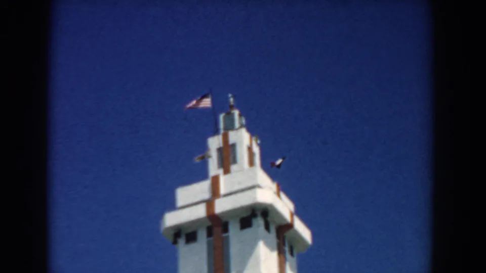 1958 home-movie frame of the tower and flags, with soft focus, grain, and strong blue sky color.