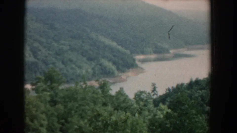 1958 South Carolina 8mm frame showing a hazy forested valley with a river bend, foreground trees, and dark vertical edges from the filmed viewpoint.