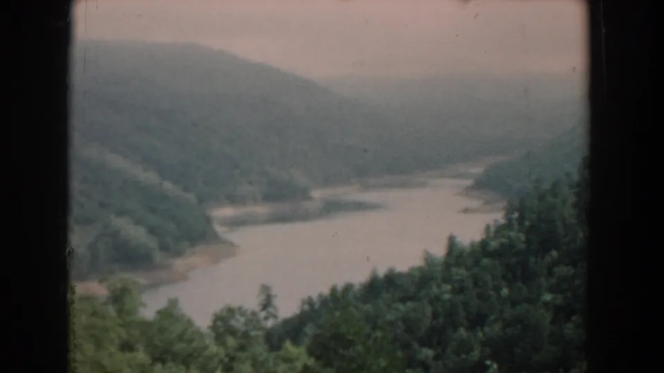 1958 South Carolina 8mm frame capturing a muted, soft-focus river valley with forested slopes, treetops in the foreground, and dark vertical borders from the original framing.