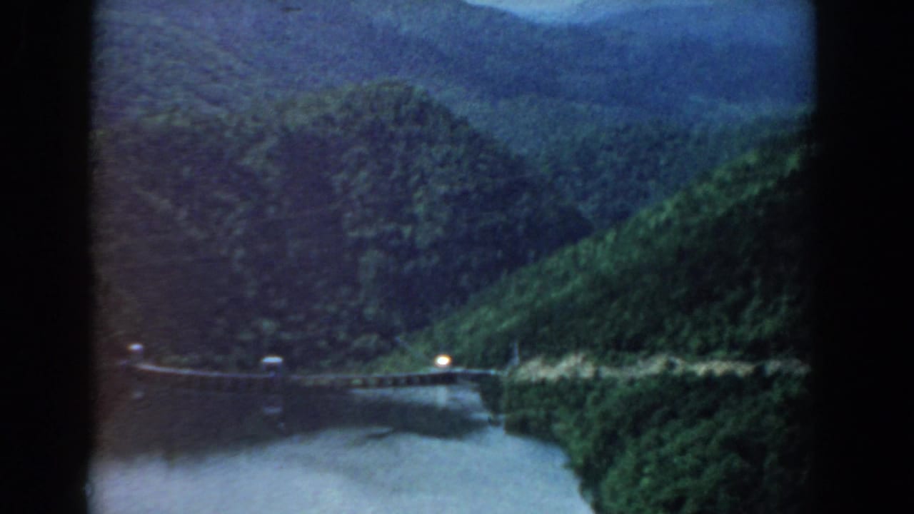 River bend below green mountain slopes with a small bridge and bright light near the shore.