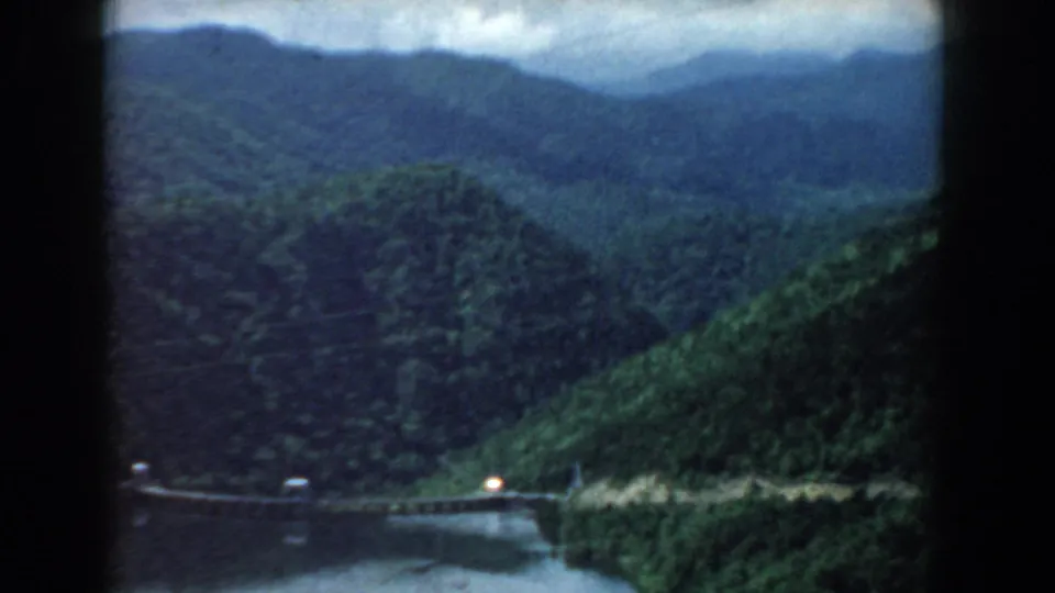South Carolina 1958 home-movie frame with layered wooded hills, river foreground, and a small bridge or road crossing at the lower edge.
