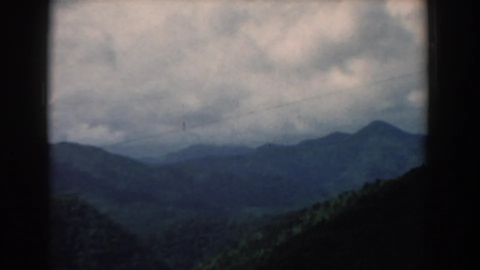 1958 South Carolina 8mm frame of rolling forested mountains under mottled clouds, with dark frame edges and visible film texture.