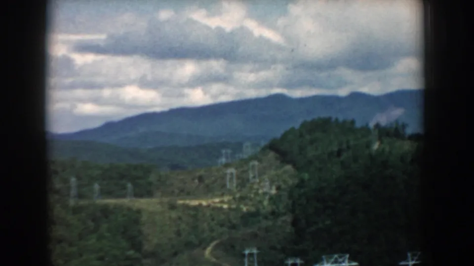 1958 South Carolina home-movie frame of a hilly forested region with a winding path and electrical towers in soft 8mm color.