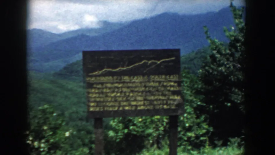 Handheld 8mm view of a dark overlook sign amid vegetation, with South Carolina mountain ridges beyond.