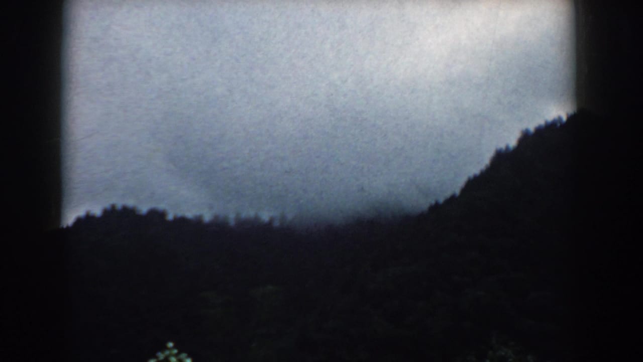 1958 South Carolina 8mm frame showing a dark forested slope silhouetted beneath low storm clouds and a pale overcast sky.
