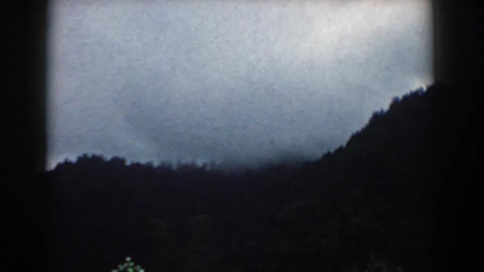 1958 South Carolina 8mm frame showing a dark forested slope silhouetted beneath low storm clouds and a pale overcast sky.