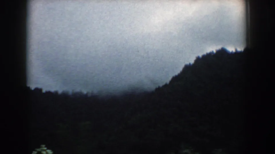 A South Carolina 8mm home-movie frame from 1958 shows a dark tree-covered ridge beneath heavy gray cloud cover.
