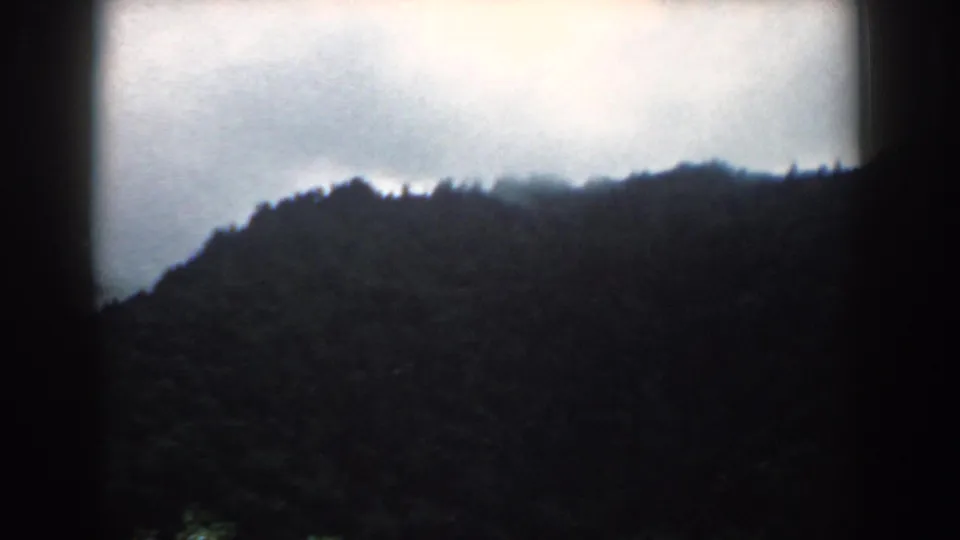 1958 South Carolina home-movie frame with a dark forested hillside silhouetted against a bright, cloudy sky.