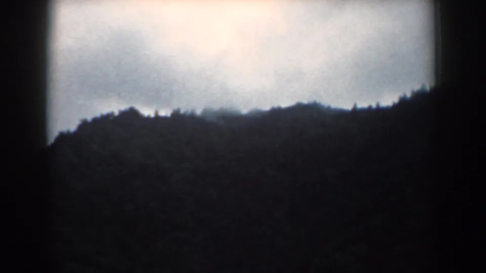 1958 South Carolina 8mm frame showing a low, shadowed hillside beneath pale cloud cover, with grain and rounded film edges.