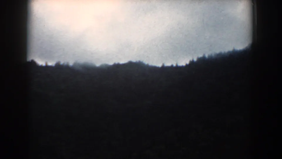 1958 South Carolina home-movie frame of a dark tree-covered ridge under a cloudy sky, framed by the natural 8mm border.