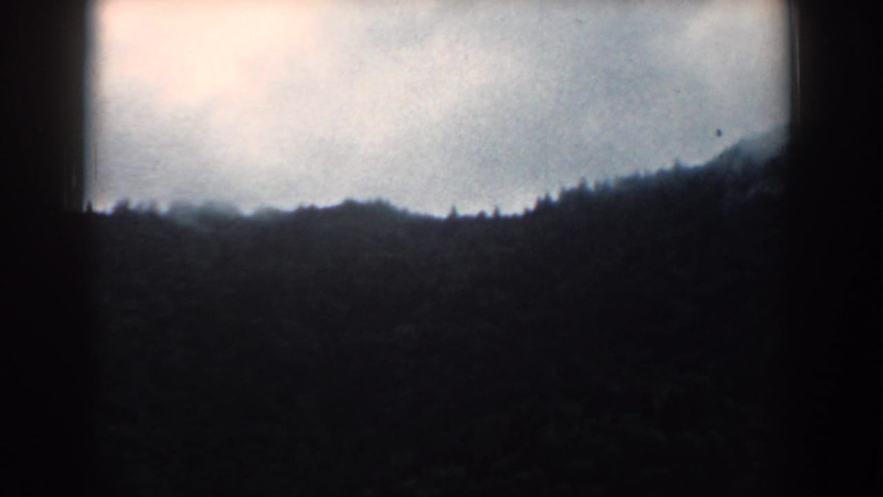 Dark wooded mountain ridge under a pale cloudy sky with heavy black frame borders.