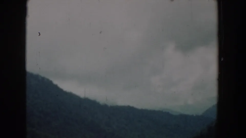 1958 South Carolina home-movie frame with a dark gate edge around a soft view of forested hills and low overcast clouds.