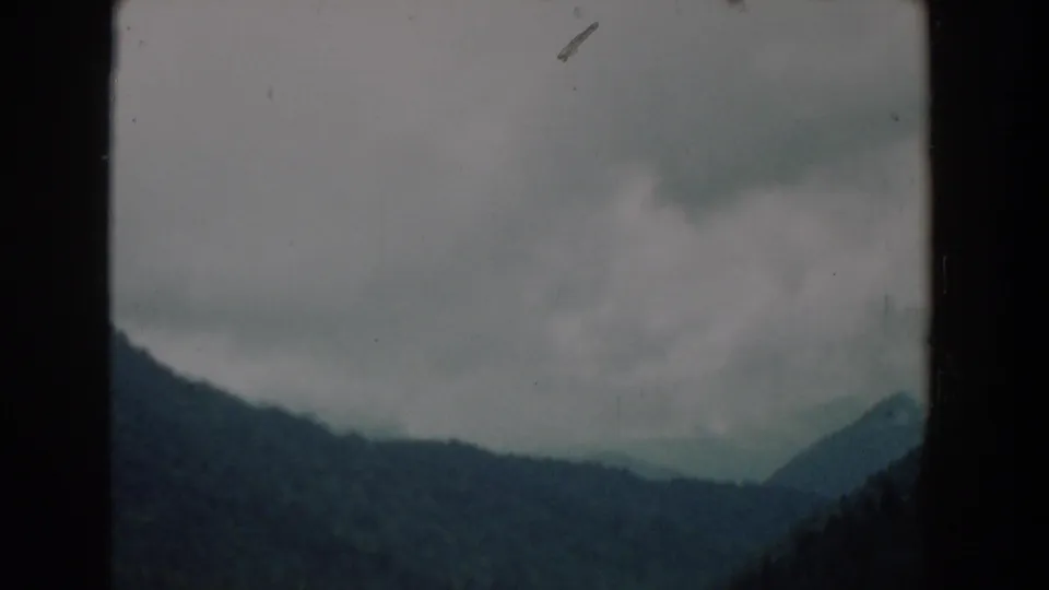1958 South Carolina 8mm frame showing layered tree-covered slopes, low cloud cover, and a distant pointed hill at the right edge.