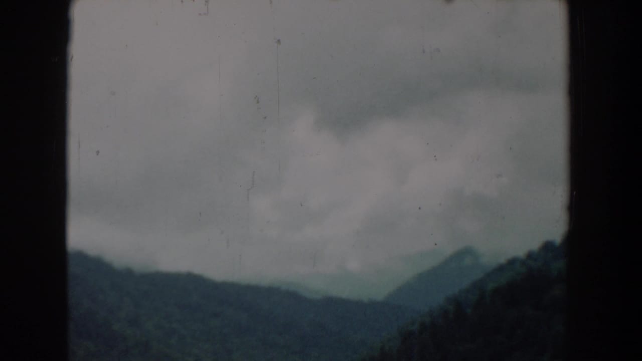 1958 8mm frame showing cloudy mountain ridges and forested slopes, likely from a moving visitor viewpoint.