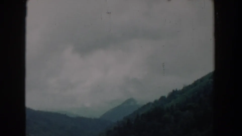 Soft 1958 8mm frame of a dark green mountain slope under low clouds, with visible film grain and border.