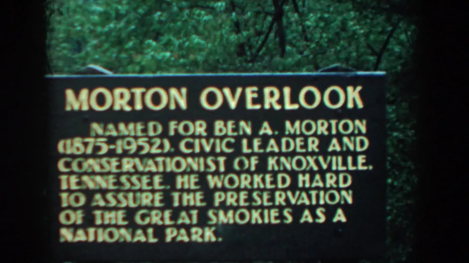 1958 8mm frame of the Morton Overlook sign honoring Ben A. Morton, seen against dense green foliage.