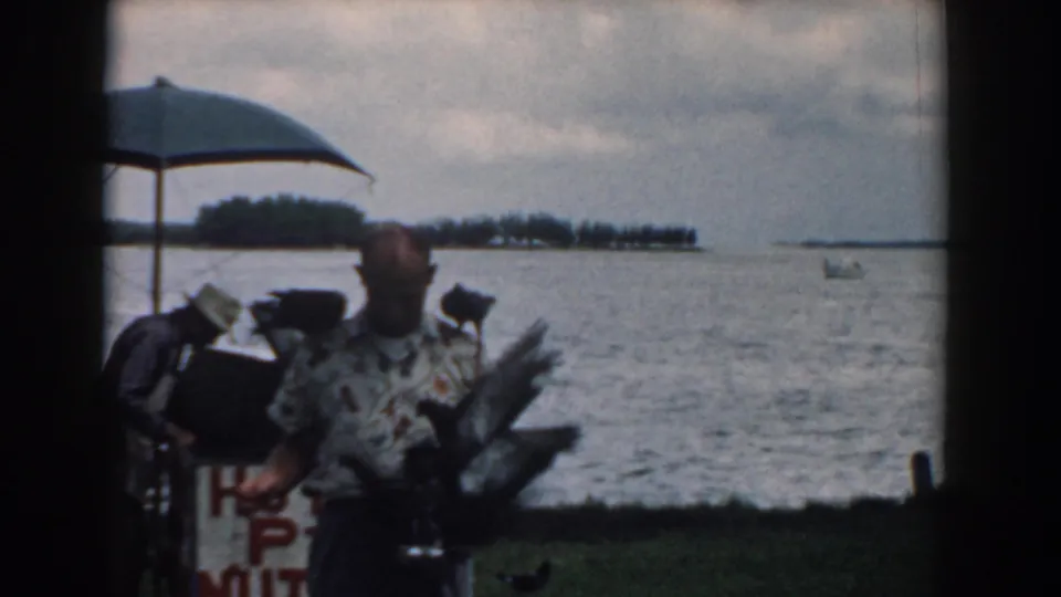 1958 South Carolina home-movie frame of a man with large birds on his shoulders near a lakeside concession stand.