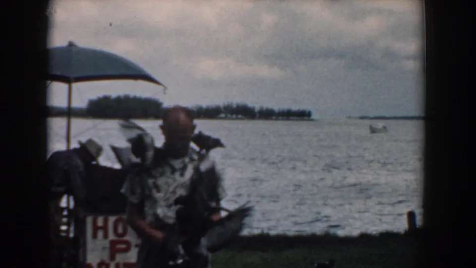 The man faces the camera with birds on his shoulders and arms beside a waterfront stand in South Carolina.