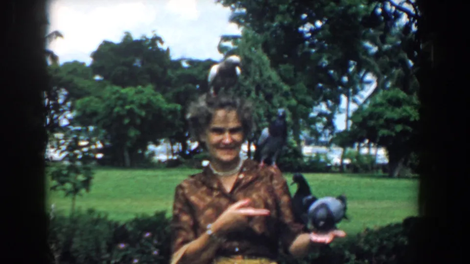 In 1958 South Carolina home-movie footage, a woman in a patterned blouse smiles while pigeons perch on her head, shoulder, and hand.