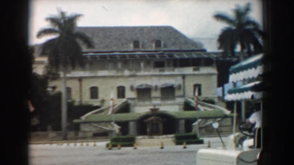 The South Carolina footage holds on the building frontage with palms, green awnings, steps, hedges, and part of another structure at the right edge.