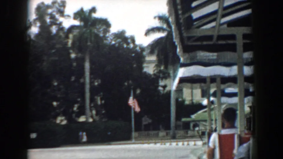 The 1958 South Carolina home-movie view shifts toward palm trees and dense landscaping, with part of the tan building and a passenger visible.