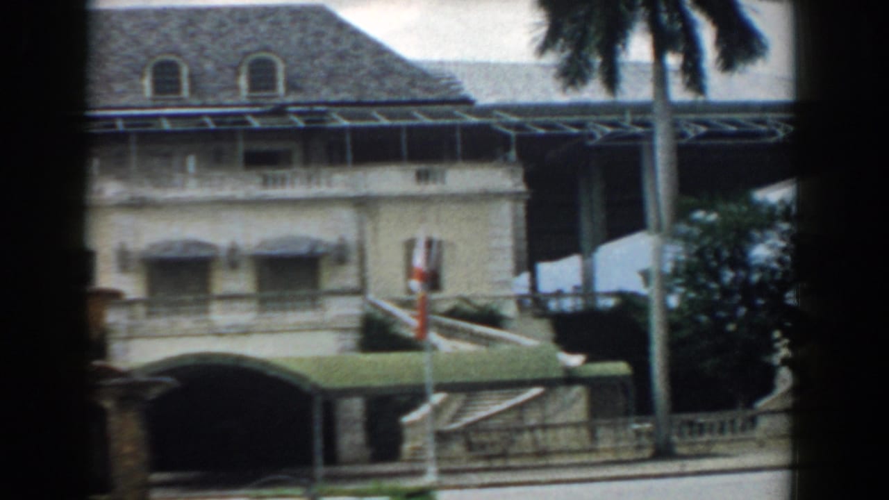 Pale multi-story building with balconies, awnings, palm trunks, and roadside posts.